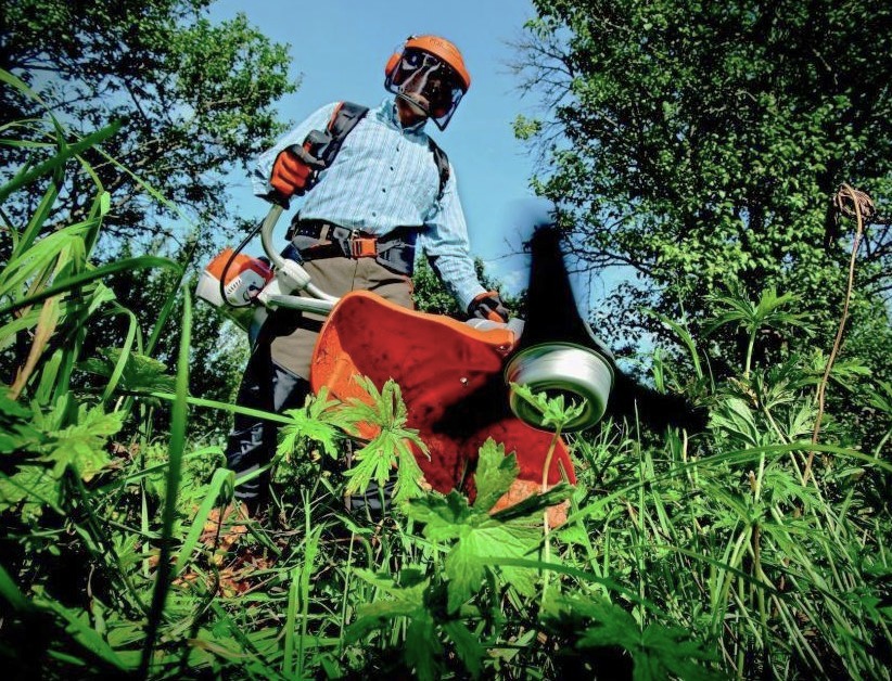 Person using a brush cutter on weeds wearing a face guard, ear protection, and gloves.