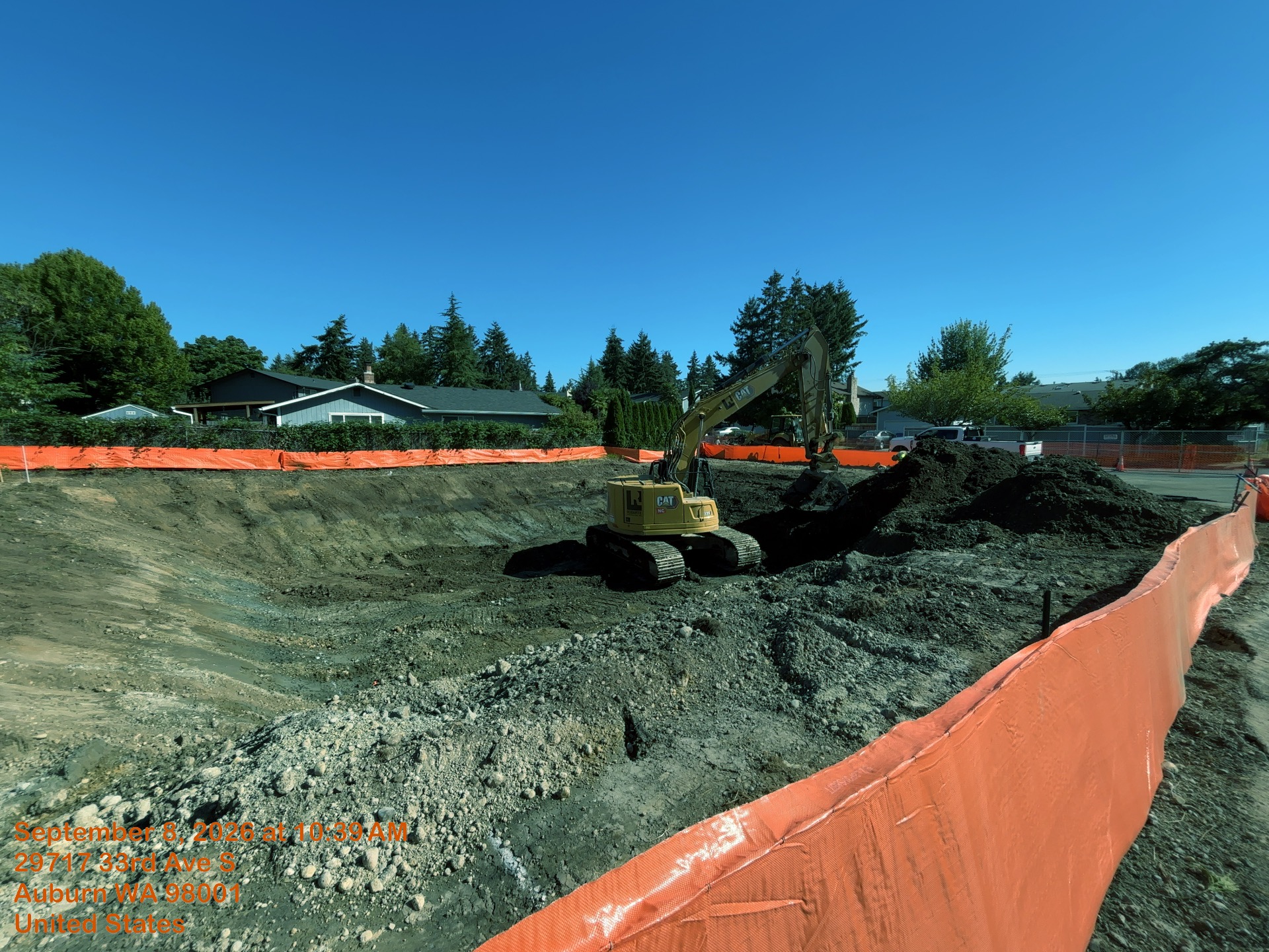 An excavator is scooping dirt out of a large sunken area that is all exposed dirt and rocks. An orange fence surrounds the sunken area. 