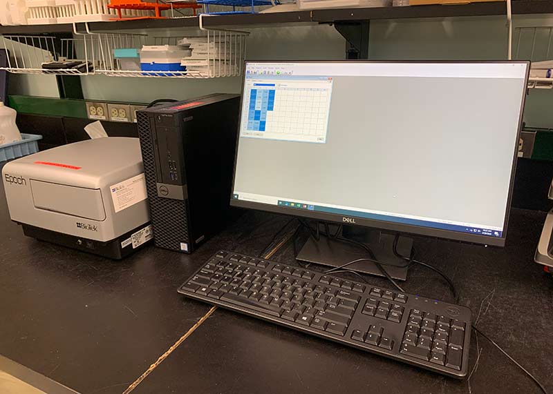 A desktop computer, monitor, and keyboard sit on a black laboratory bench next to a grey Spectrophotometer. The screen shows a grid with boxes of varying shades of blue. This instrument is used to measure algal toxins in water samples.  