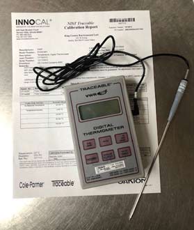 A grey and red digital reference thermometer and calibration certificate on a stainless-steel laboratory countertop.