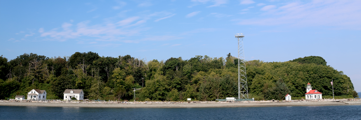 A view of Vashon's Point Robinson Lighthouse and adjacent buildings on the east side of the Island. A small red and white light house stands beside a tall radio tower. Three white buildings stand further down the beach. Green trees and blue sky frame the scene.