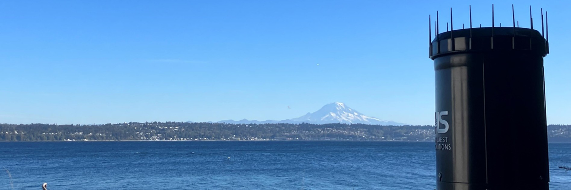 A round black rain gage with blue water, blue sky, and Mount Rainier in the distance
