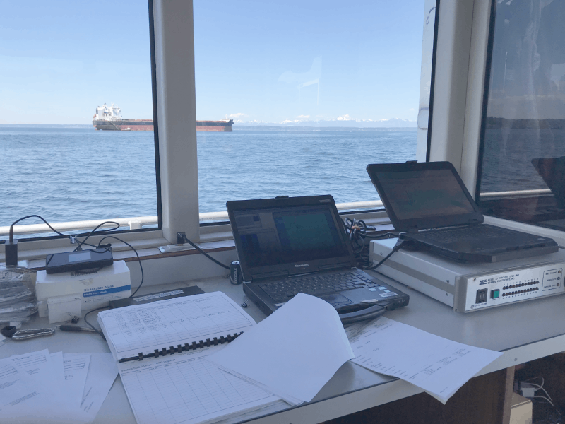 Photo of electronic sampling equipment in the cabin of a research vessel, with water and a barge in the  background.