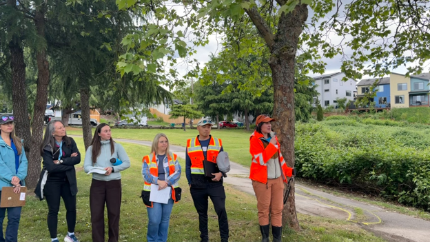 Five people standing next to a person with a high visibility jacket who is speaking. There are trees, grass, and shrubs around the people and a paved trail goes through the grassy area. Some hones can be seen in the far background.