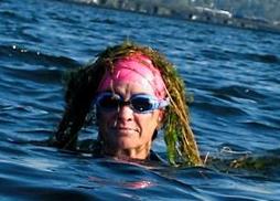 A swimmer in goggles poking their head out of a lake. Milfoil weed is draped over their head like long hair.