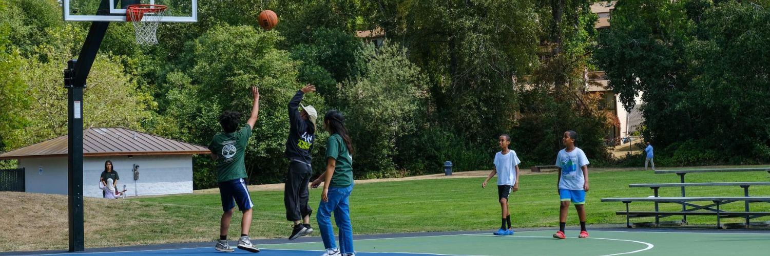 5 people playing basketball on a basketball court with the ball in the air. In the background is a grassy field with building and bleachers. A child rides a scooter with adults walking on the grass.