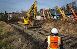 A backhoe travels down a rail-bed with a worker watching