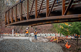 A steel bridge is being lowered over the Black River with two construction workers in the distance
