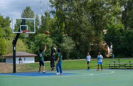 Five people play basketball on a basketball court, the ball is in the air. A grassy field with a building is in the background with a child riding a scooter and adults walking by, 