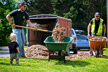 Two volunteers putting wood chips in wheelbarrows with a toddler watching
