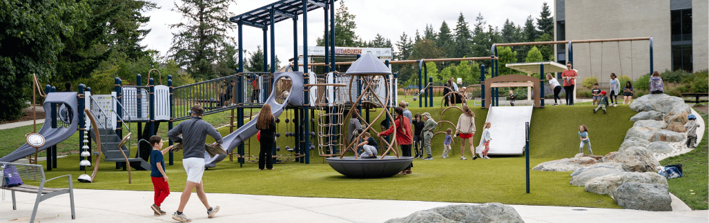 A wide image of a playground with many children playing on it. There is green grass in the foreground and a forest in the background on a cloudy day.