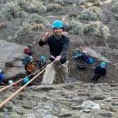 A young man in a blue helmet makes a peace sign and smiles to the camera. He is rock climbing and suspended in the air on a rope.