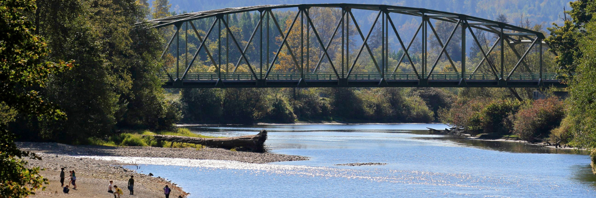 People relax on a river bank beneath a bridge