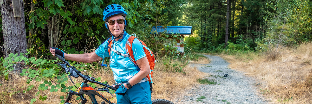 A woman stands with her bike at the entrance to a gravel trail going into the woods