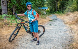 A woman stands with her mountain bike at the entrance to a wooded gravel trail.