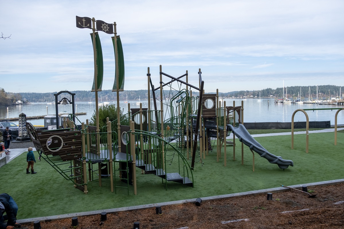 A green and brown playground shaped like a ship. A marina with boats is in the background