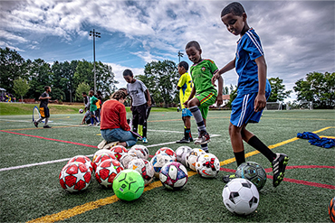 A group of kids come up to a pile of soccer balls on a turfed field. Other kids are playing soccer in the background.