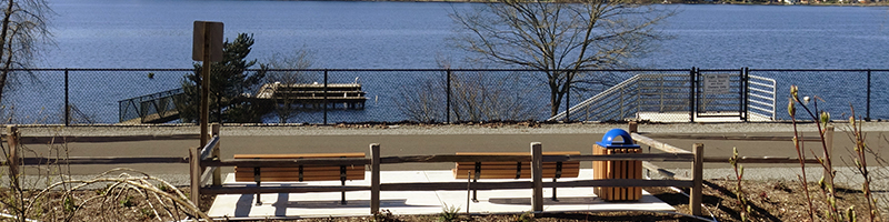 A view of a rest area with two benches and a trashcan on the trail with the lake in background