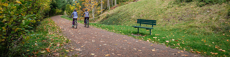 Two bikers ride away along the gravel trail with a green bench just off the trail