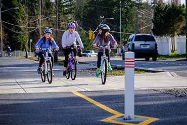 3 kids ride bicycles on a wide, paved trail with a bollard in the foreground.