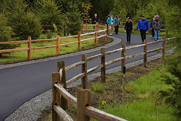 People stroll along a paved path lined with conifers and a split-rail fence.