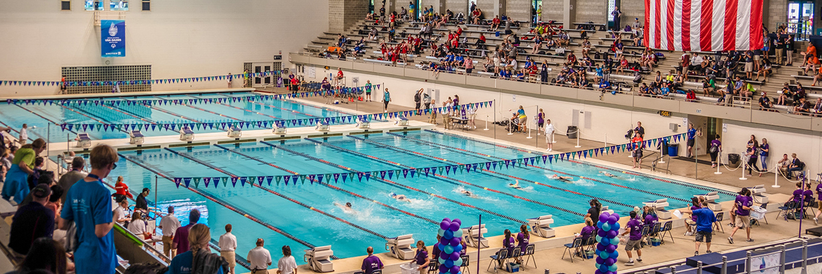 people swimming in an olympic-size lap pool with others watching from the bleachers