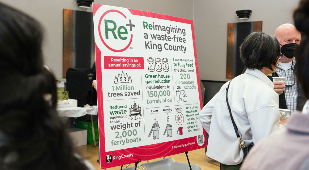Image showing several people visiting at a public meeting. A "Re+ Zero Waste" display stands in the center of the room.
