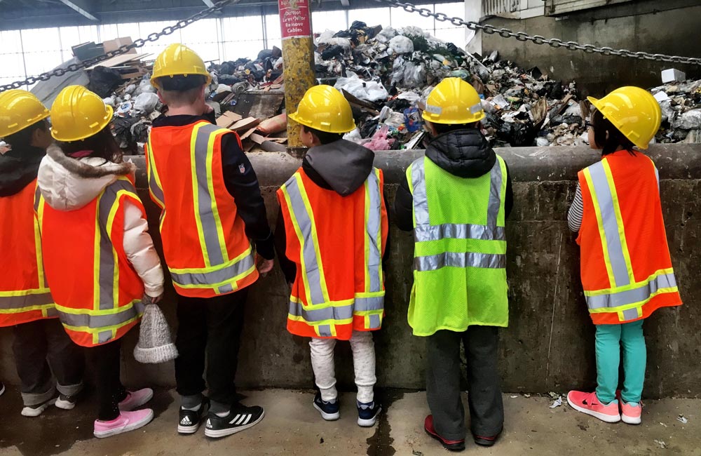 Six students wear reflective vests and yellow hard hats while observing a recycling facility.