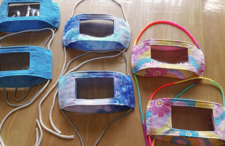 Image showing six window masks (or smile masks) on a wooden table. Window masks are reusable cloth face masks with a clear plastic panel, which can help people communicate with those who are deaf or hard of hearing.