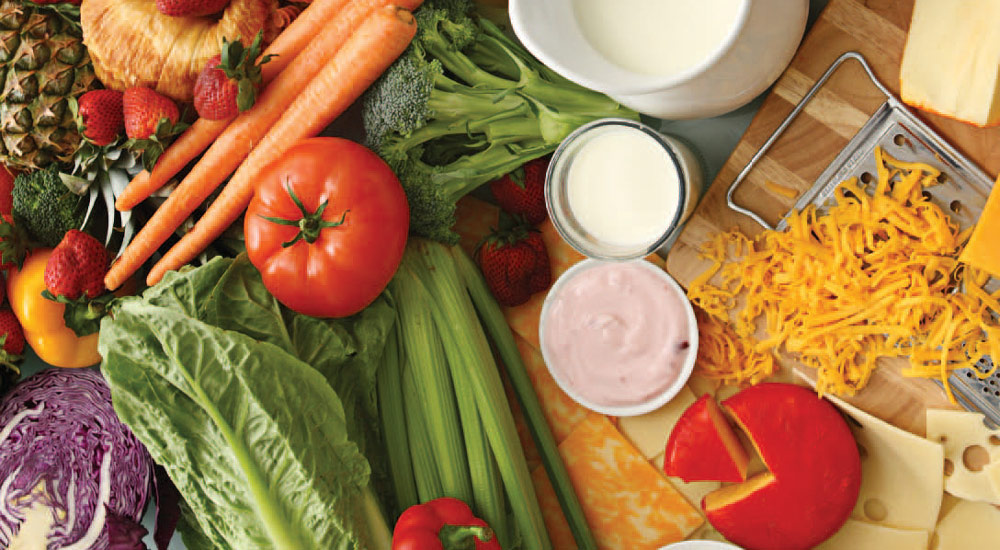 An image of fresh vegetables, cheese, and a bowl of salad dressing on a wooden countertop.