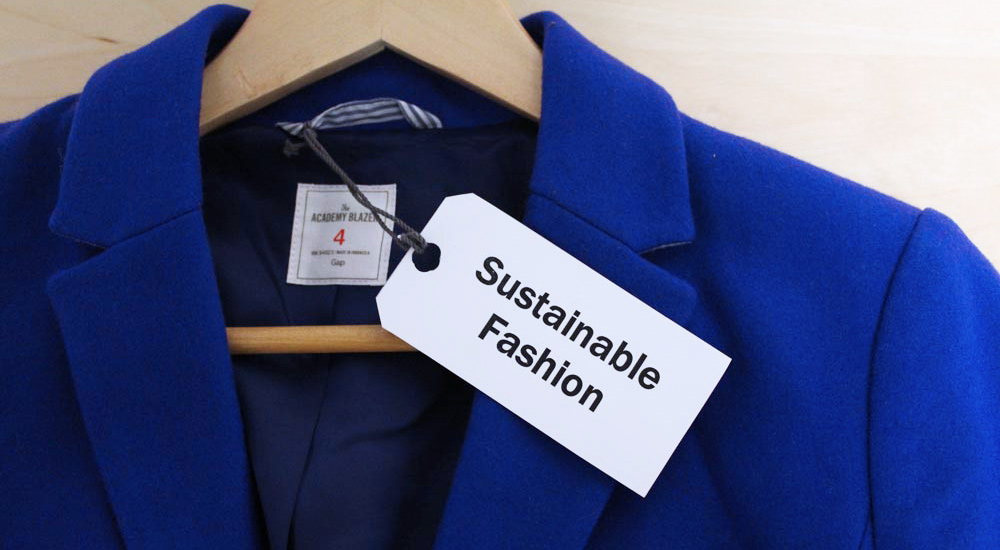 An image of a navy blue jacket or blazer on a hanger. An attached white tag says Sustainable Fashion.