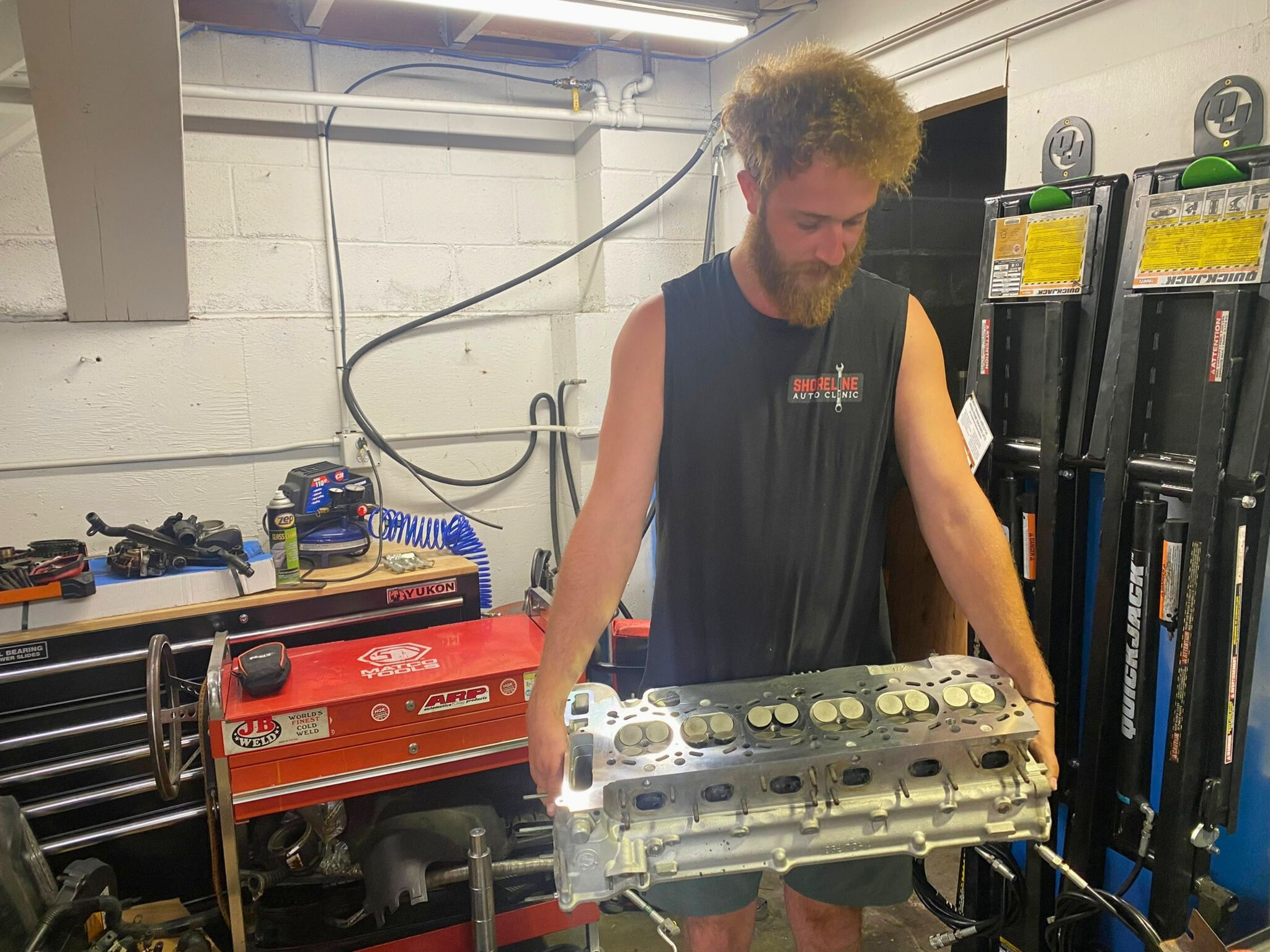 A man in a sleeveless black T-shirt holds a heavy, shiny metal automotive part in an auto shop.