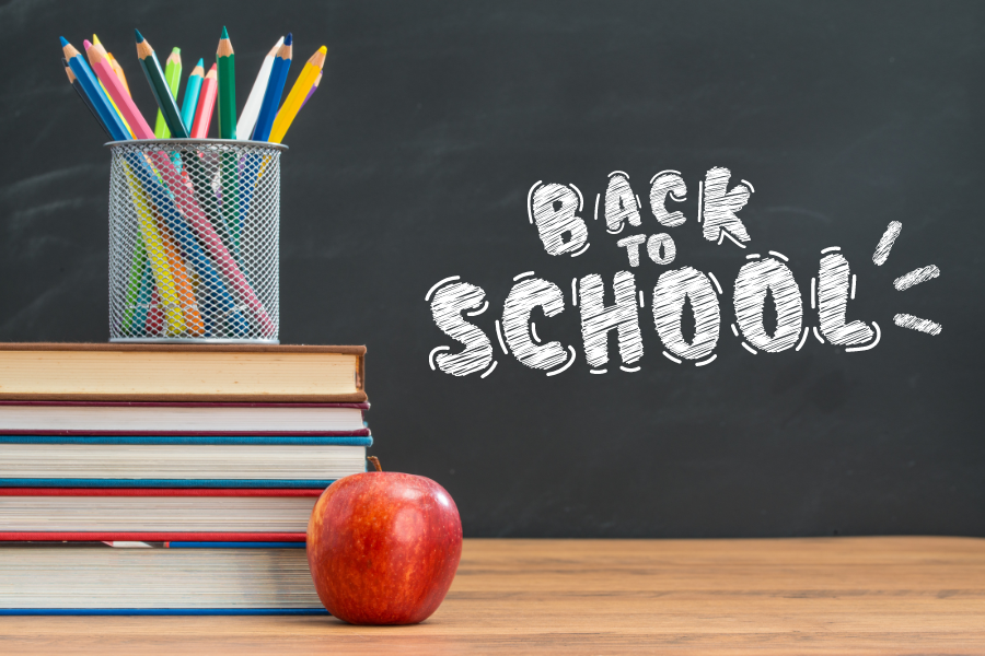 A stack of books, pens in a cupholder, and a red apple sit in front of a chalkboard that says "Back to School"