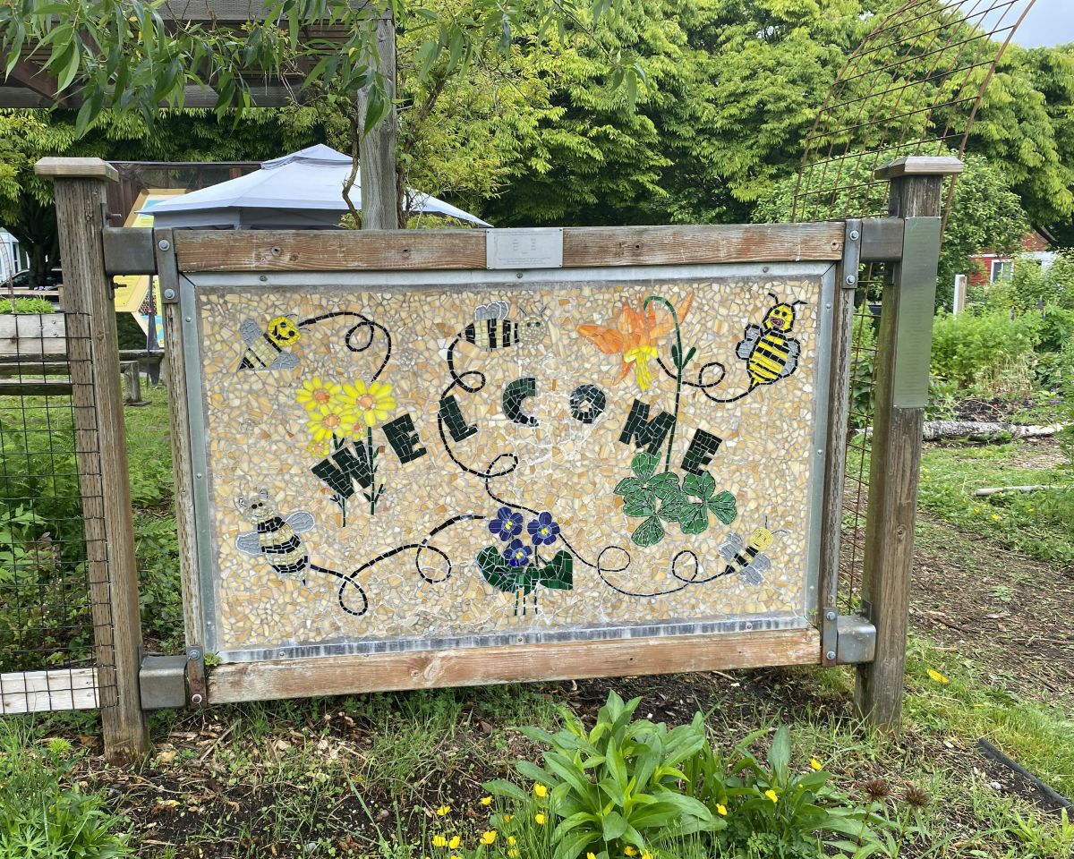 A mosaic art sign with illustrations of flowers and bees is framed by a wooden fence at the entrance of a garden.