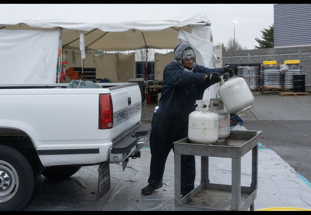 A collection worker lifts a white propne tank from a truck bed to a cart with other propane tanks
