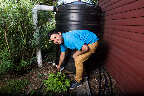 A man in a blue T-shirt checks a valve on a large black water cistern by the side of a red house.