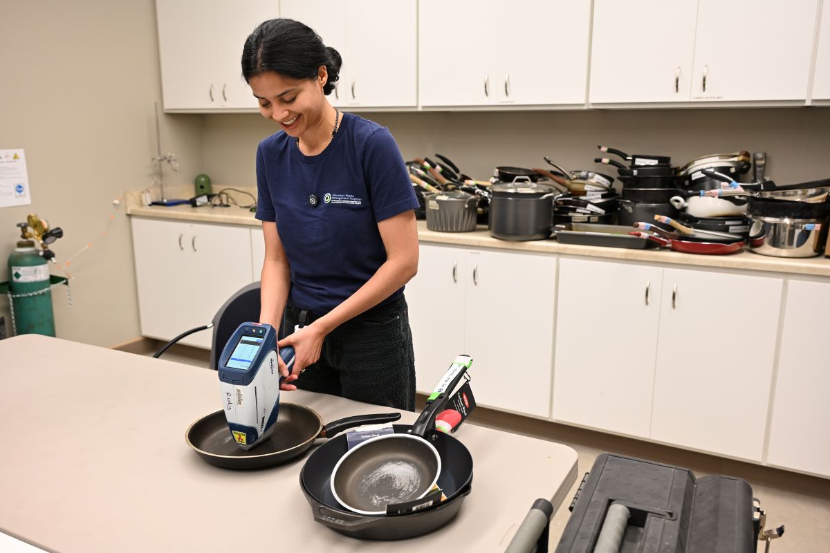 A woman in a blue T-shirt aims an XRF analyzer tool at a cooking skillet on a white table. Behind her are piles of cooking pots on a countertop with white cabinets above and below.