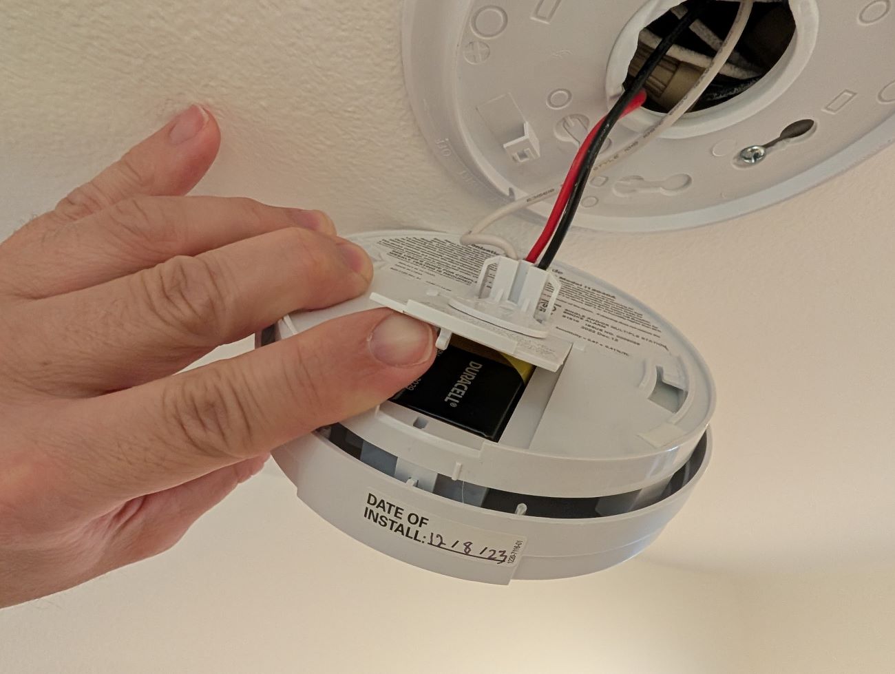 A person's hand holds a fire alarm attached to a ceiling and shows the battery compartment of the fire alarm for replacing batteries