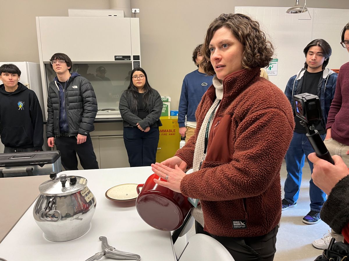 A woman holding a red cooking pot speaks to high school students in a science lab.