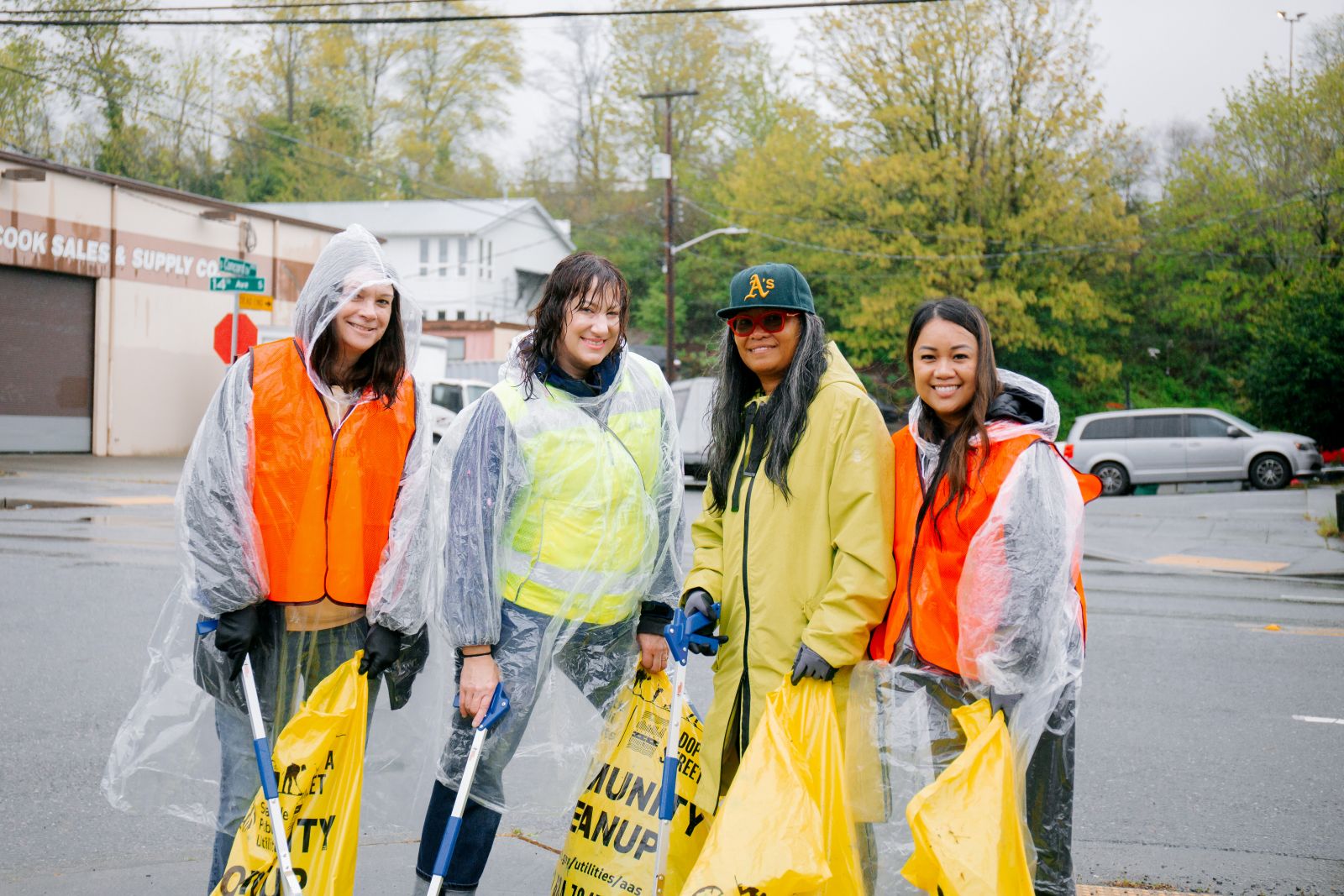 Four people in rain ponchos hold trash bags and pose for a photo in a rainy parking lot.