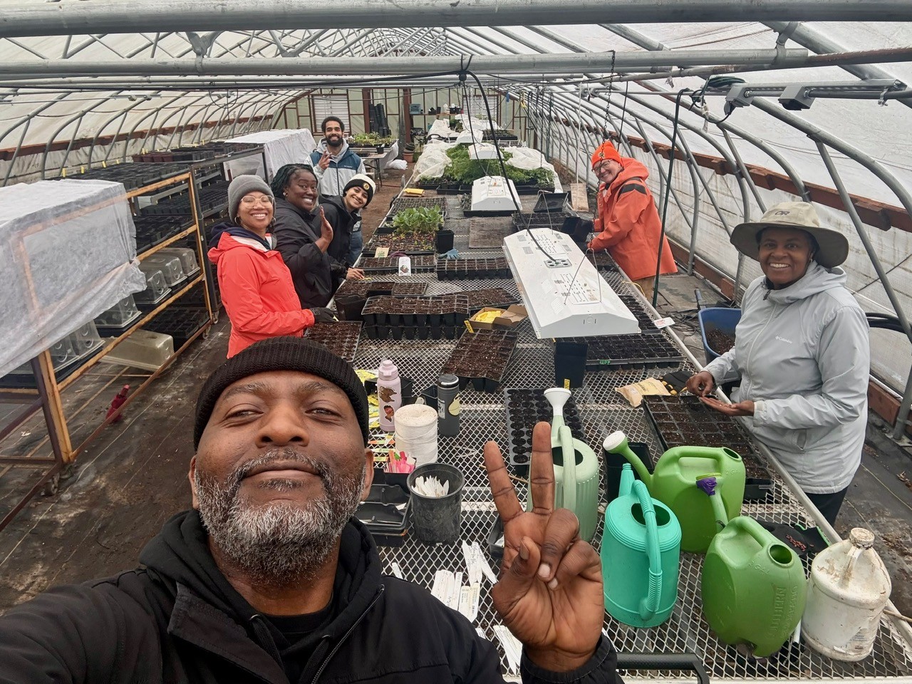 People pose for a selfie in a long green house. A long, metal grated table with watering cans and seedling pots is in the middle.