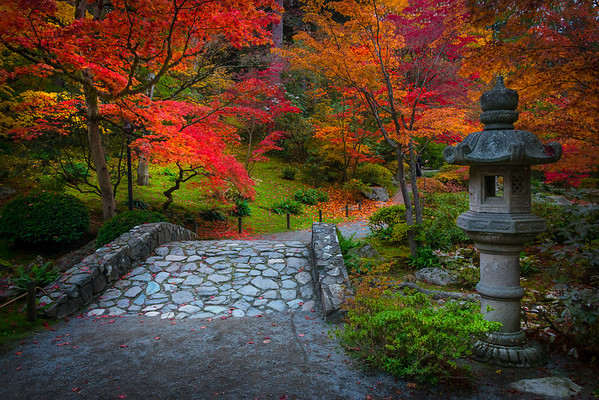 A stone path leads into a hillside with trees with red and orange leaf foliage and a small stone statue stands on the right