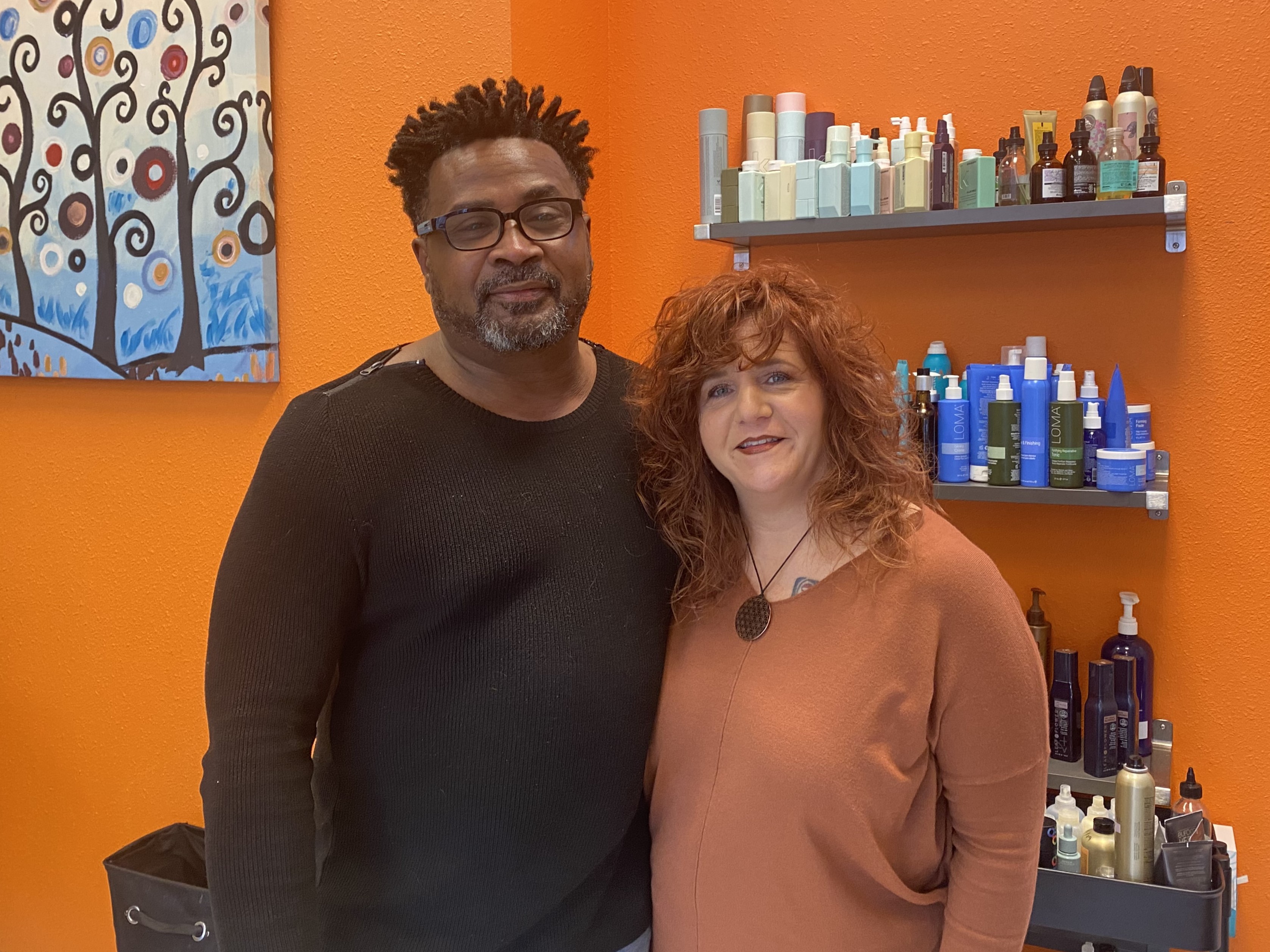 Emmett and Jessica Henley pose in front of an orange wall with shelves full of hair care products.