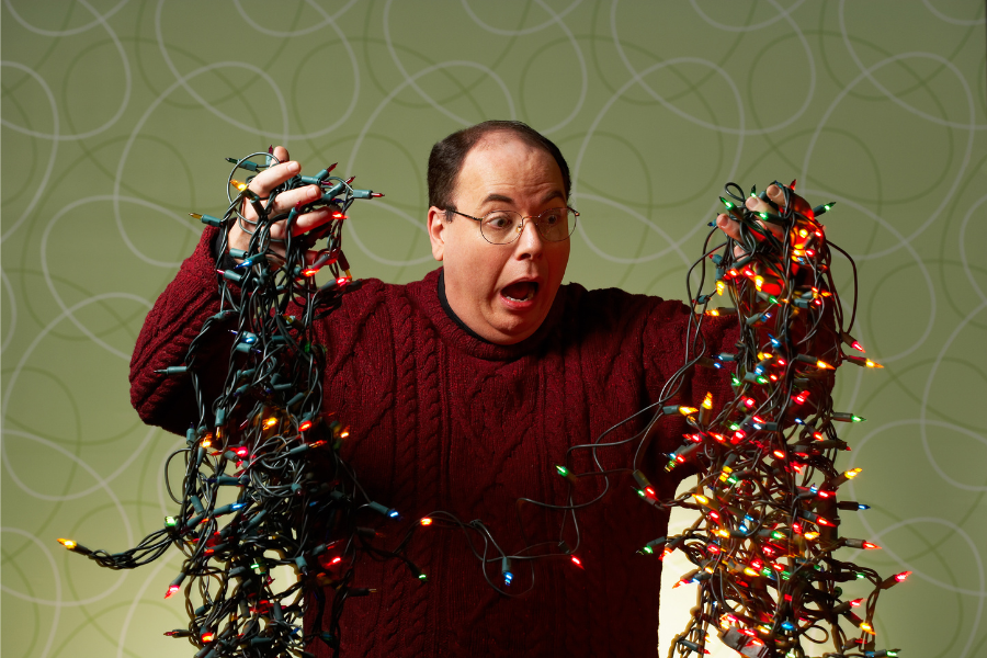 A man in a red sweater holds tangled holiday lights in both hands, looking exasperated.