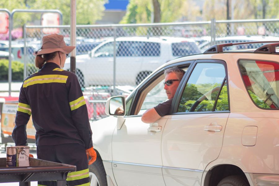 A man driving a white car speaks out a car window to a Wastemobile employee wearing a worksuit with yellow reflective colors and a hat