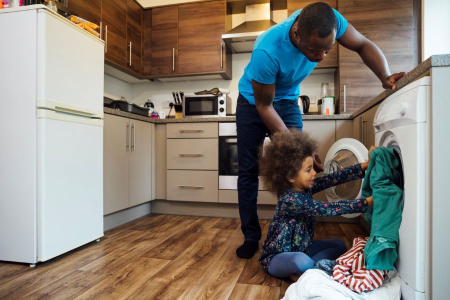 An image of man in a blue shirt holding a washing machine open while a toddler helps load laundry into the machine in an apartment.