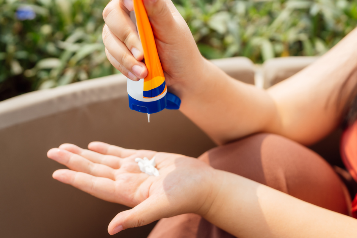 A person squeezes a white dollop of sunscreen out of an orange tube onto their left hand.