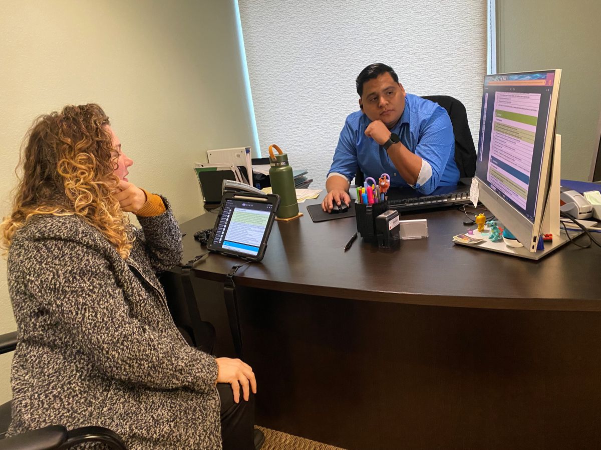 A woman and a man sit at opposite sides of a desk and look at a computer screen.
