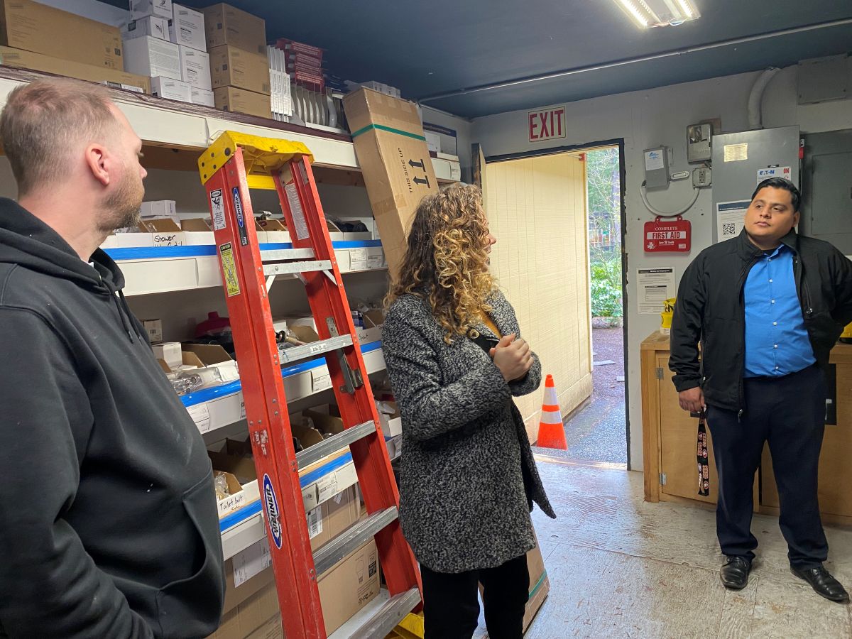 People stand and talk inside a utility shed. In the background, a folded orange ladder leans against shelves of supplies.