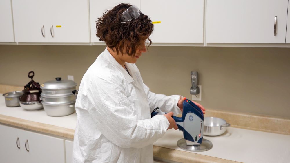 A scientist wearing a white lab coat uses an x-ray fluorescent analyzer to test a cooking pot lid for the presence of heavy metals.